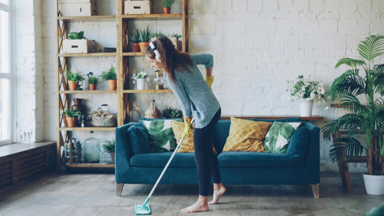 A woman wearing headphones mops a tidy living room with a blue sofa and plants, reflecting All Hours Air’s reliable, 24/7 service.
