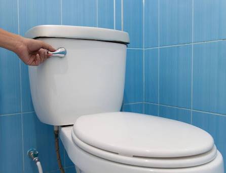 A hand presses a toilet flush handle on a white toilet against blue tile—All Hours Air ensures prompt, reliable help whenever you need it.