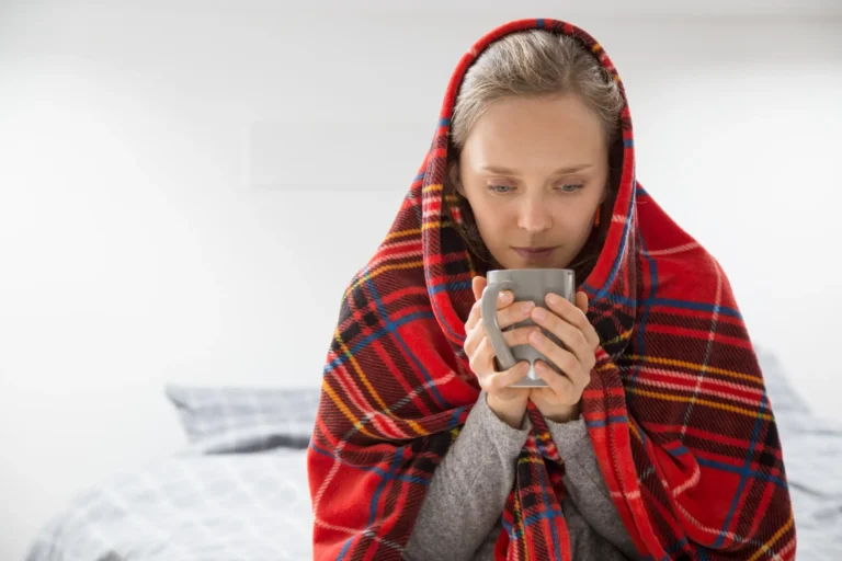 A young woman shivering under a bright red tartan blanket while holding a mug of hot cocoa, illustrating how a furnace without an annual tune-up can fail to keep a home warm.